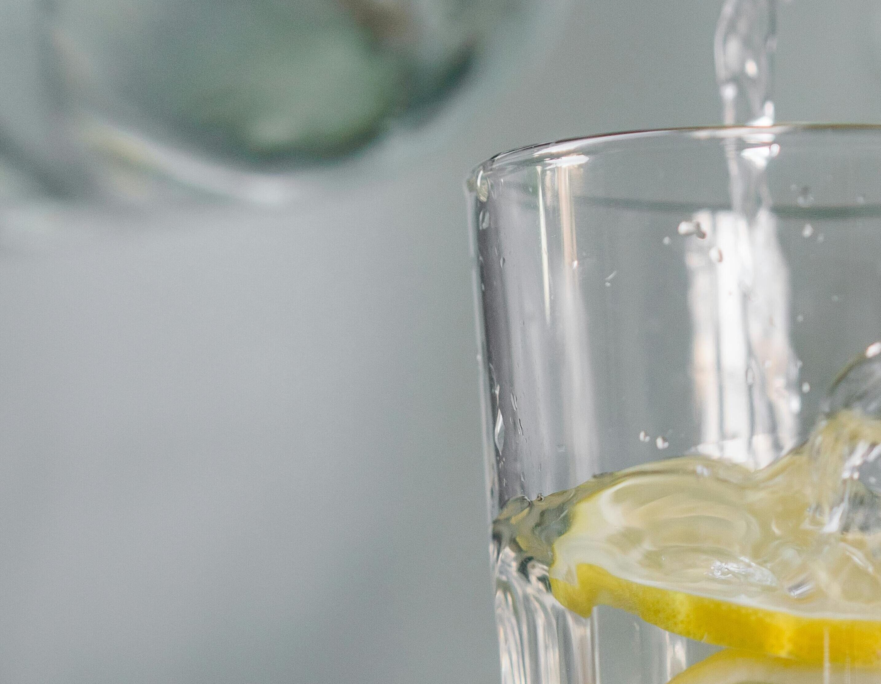Somebody pouring water from a glass bottle into a glass tumbler filled with water and lemon slices